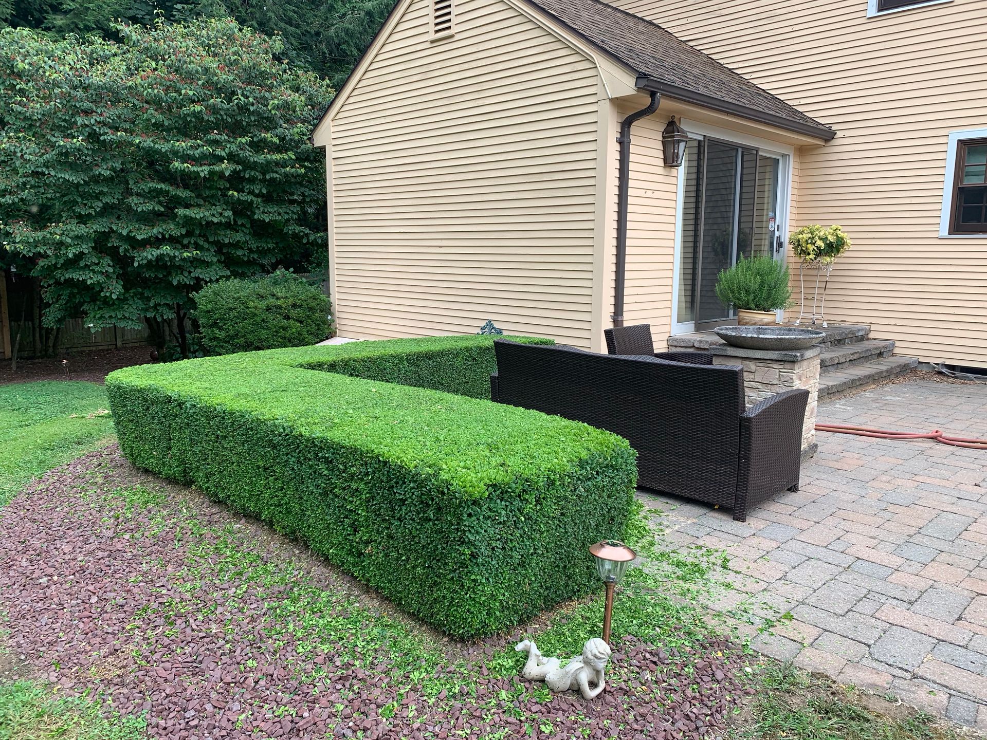 L-shaped green hedge on a brick patio beside a beige house, with outdoor seating and a small statue in front.