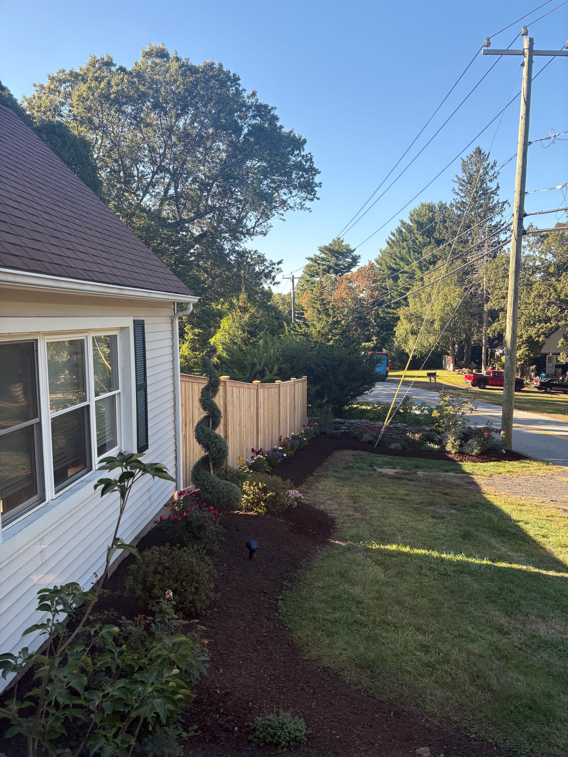 A house with a white exterior and a new wooden fence. Green lawn and trees with a blue sky.