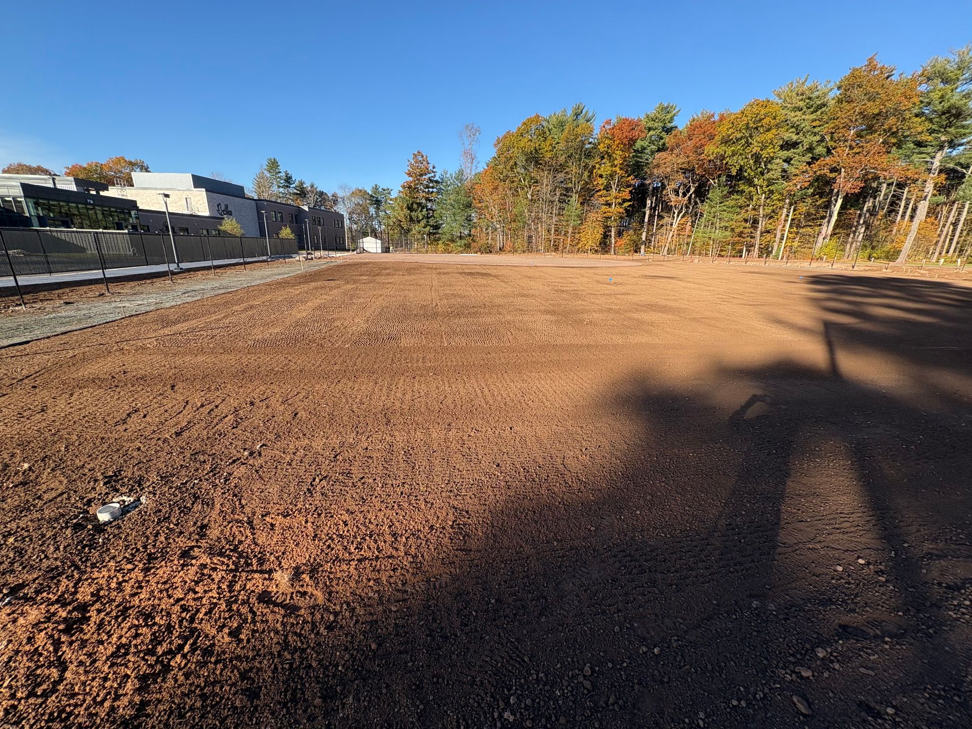 An open, cleared dirt field with trees and buildings in the background under a blue sky.