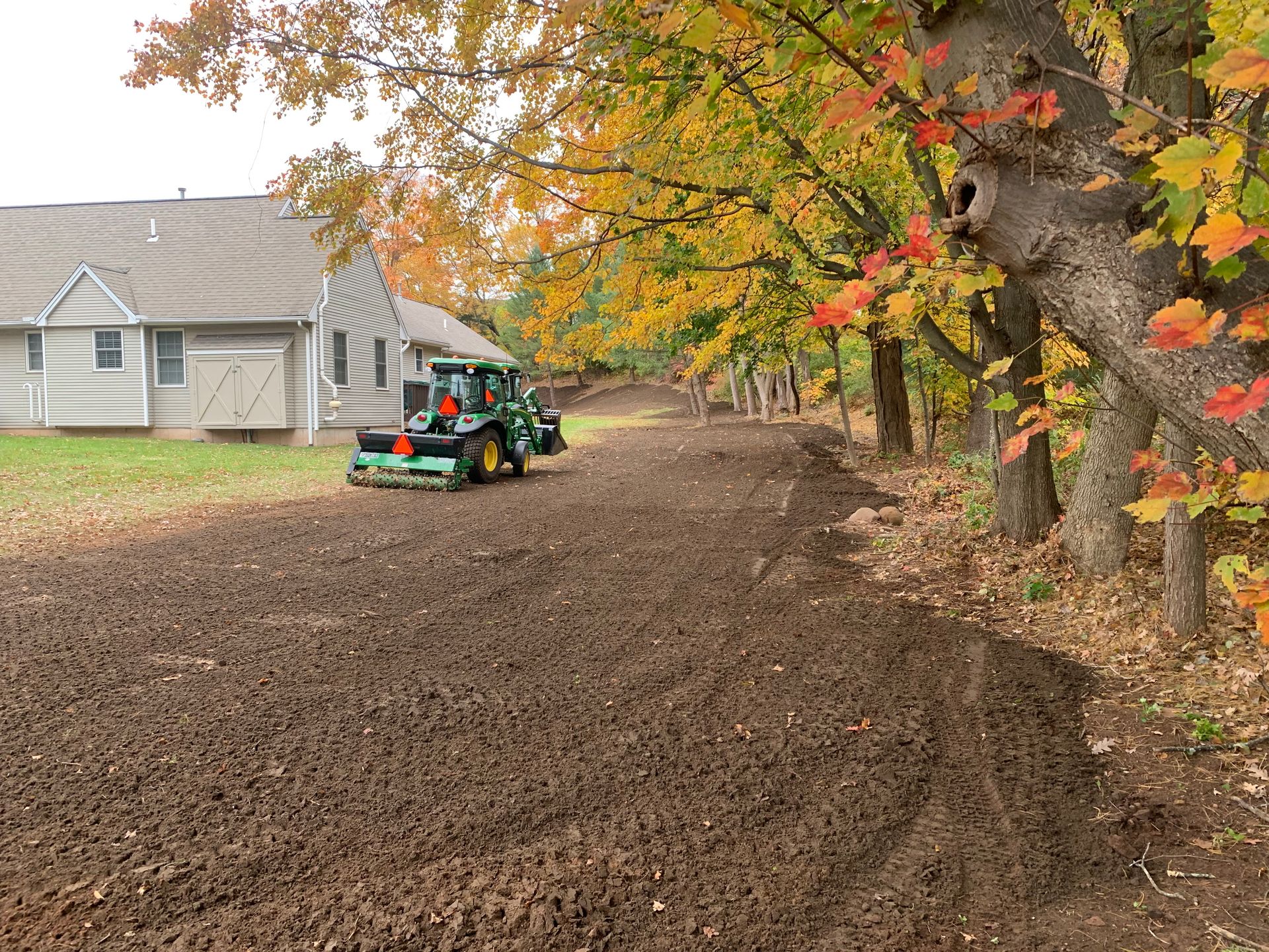 Tractor tilling soil next to a building and trees with autumn foliage.