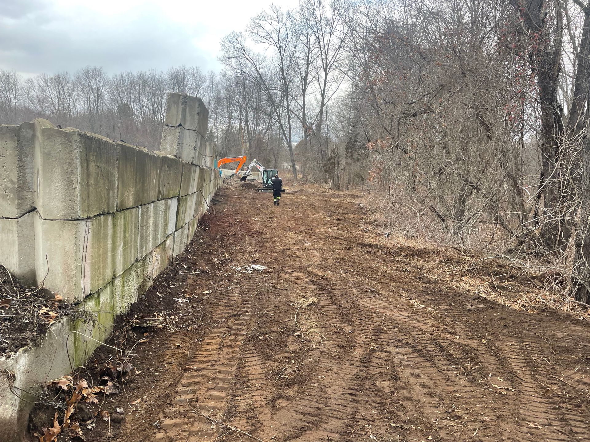 Muddy path next to a concrete block wall and trees; construction equipment visible in the distance.