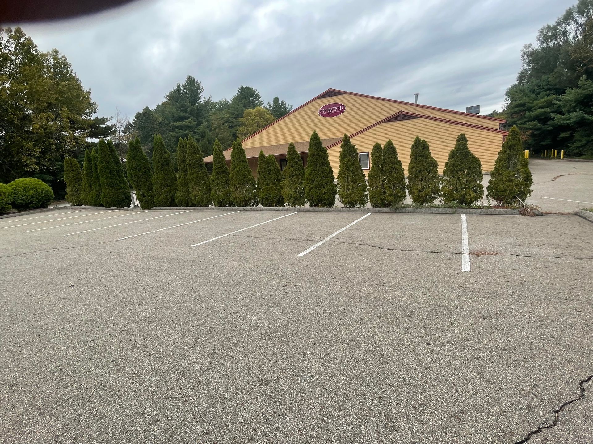 Restaurant with tan roof and row of green trees in front, in a gray parking lot under a cloudy sky.