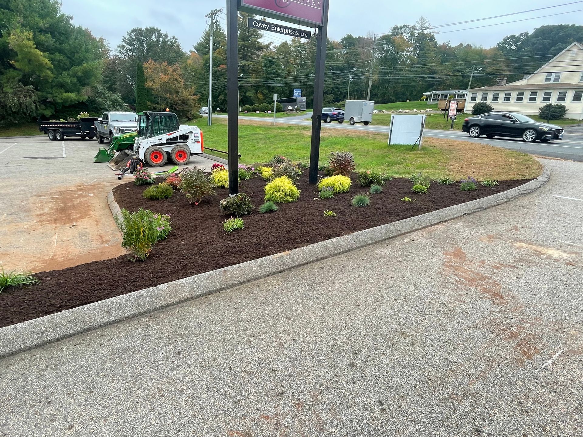 Landscaped flower bed with mulch edging a gravel driveway, with a Bobcat, truck, and car.
