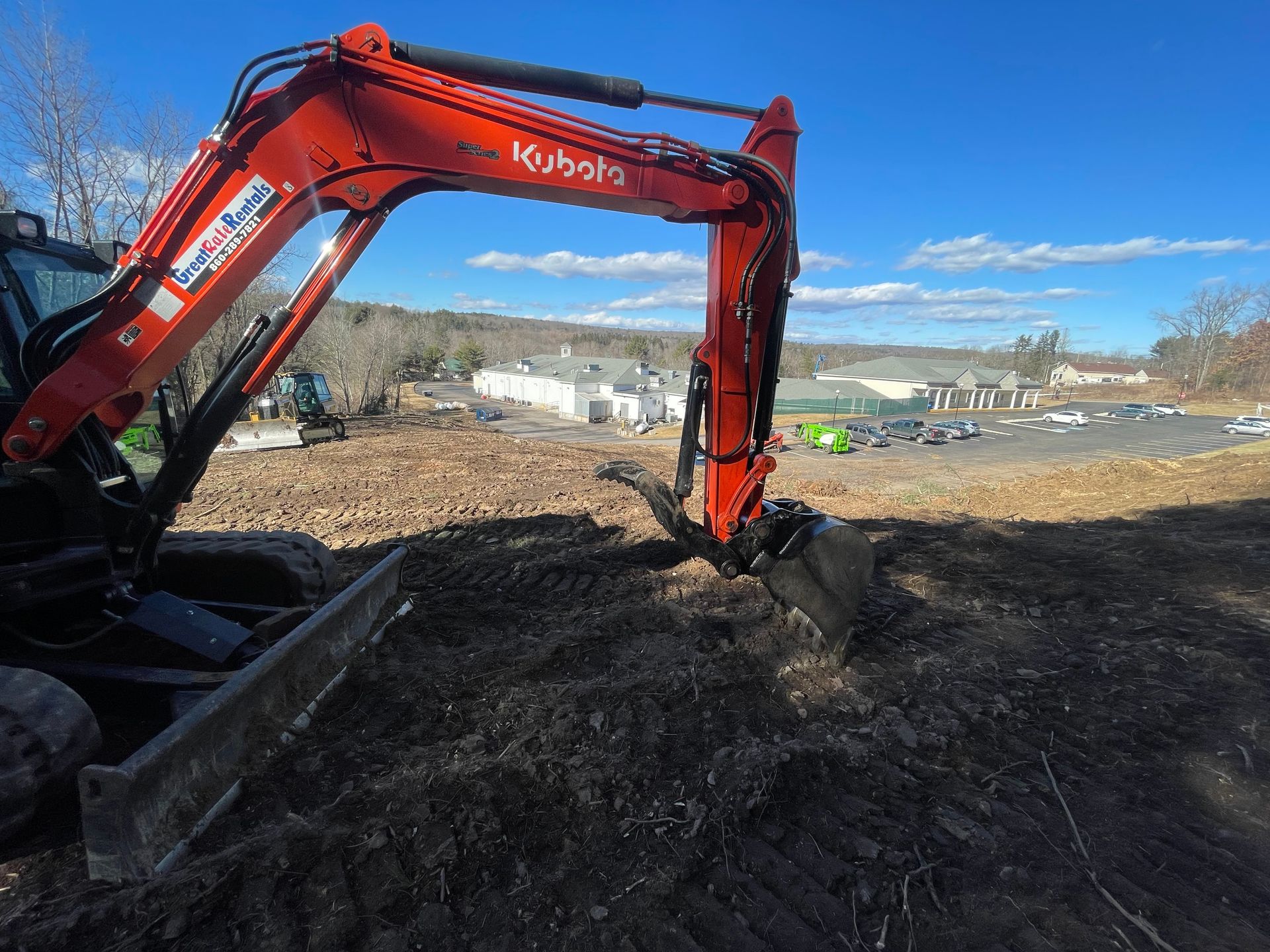 Red Kubota excavator digging in a field on a sunny day. Background includes buildings and trees.