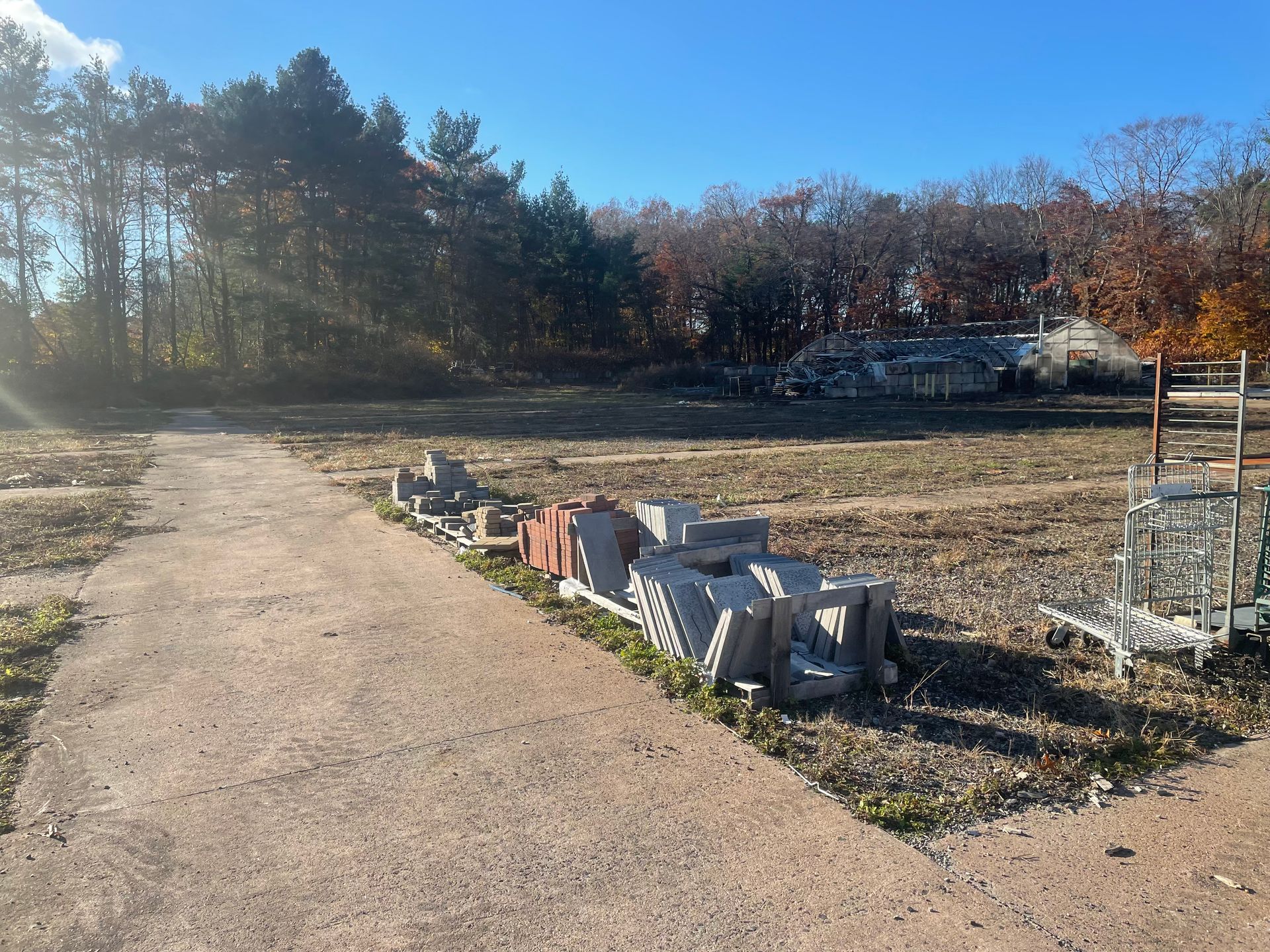 Construction site with concrete pathway, bricks, and equipment against a backdrop of trees under a blue sky.