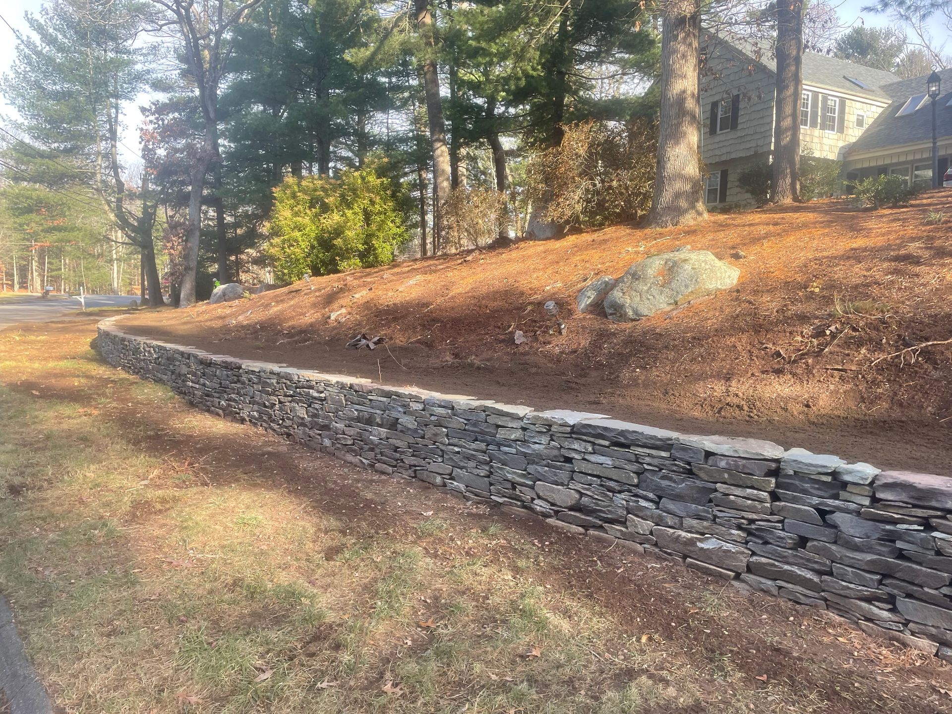 Stone retaining wall along a sloped, mulched bank with trees and houses in the background.
