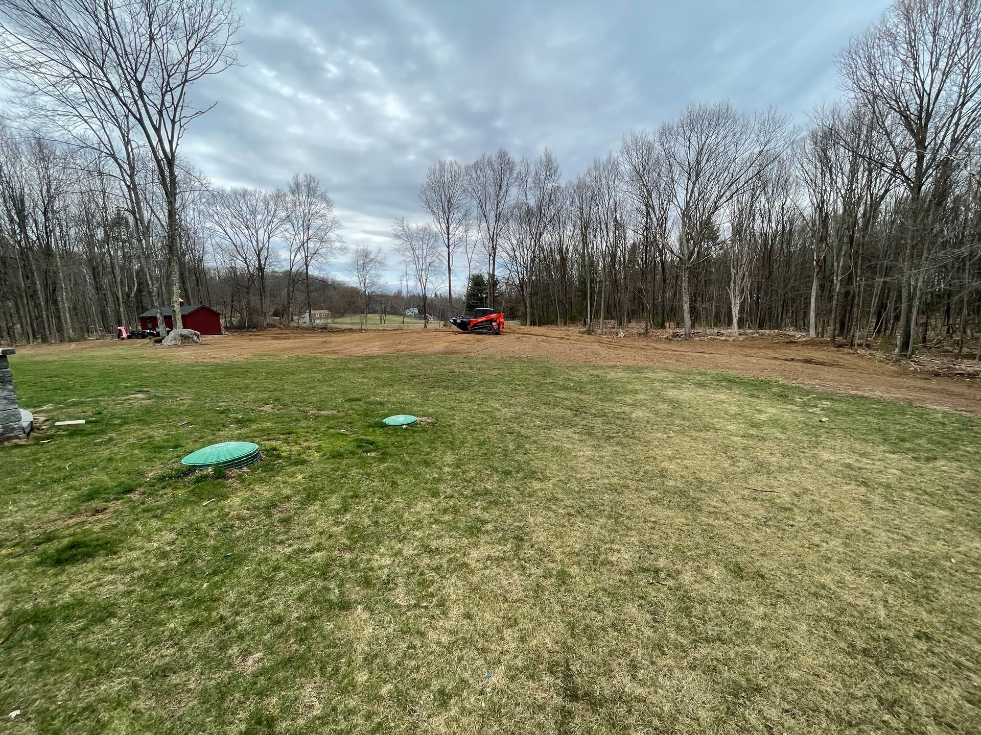 Grassy field with bare trees in the background under a cloudy sky. Red shed and small tractor.