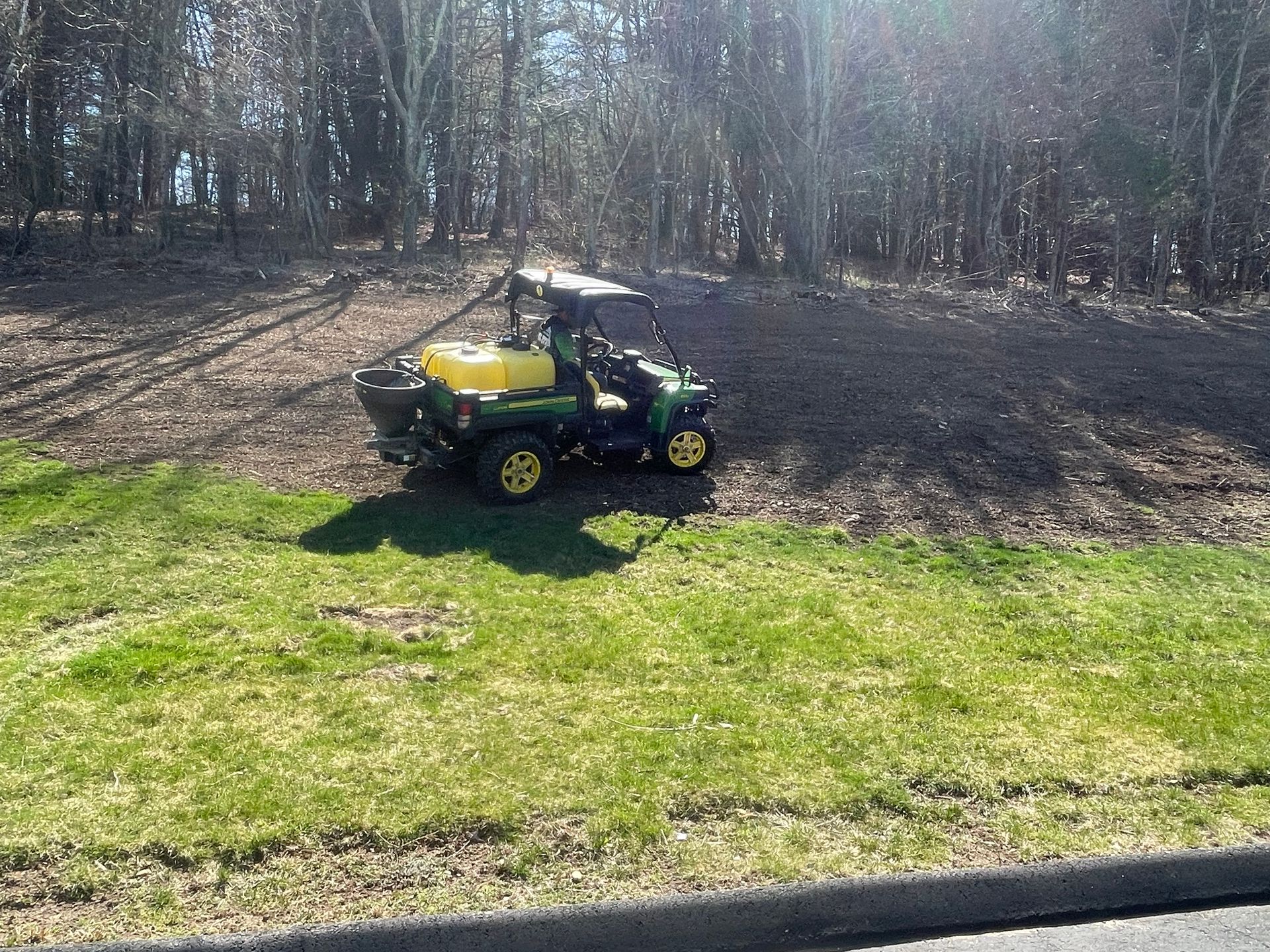 Green utility vehicle with yellow tank spreading material on a hillside next to a wooded area.