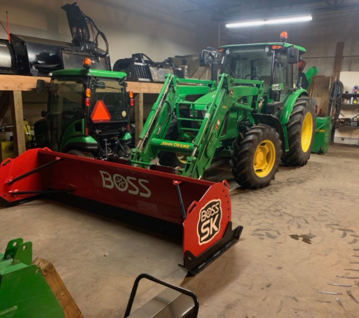A John Deere tractor with a front-mounted red BOSS snowplow inside a garage. Another tractor is in the background.