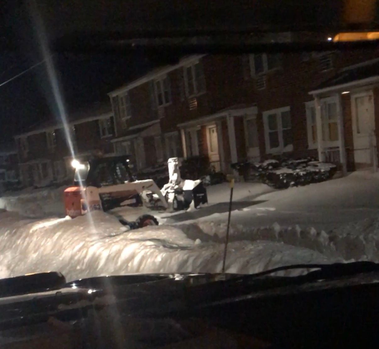 Snow-covered street at night; snow blower clearing paths in front of houses.