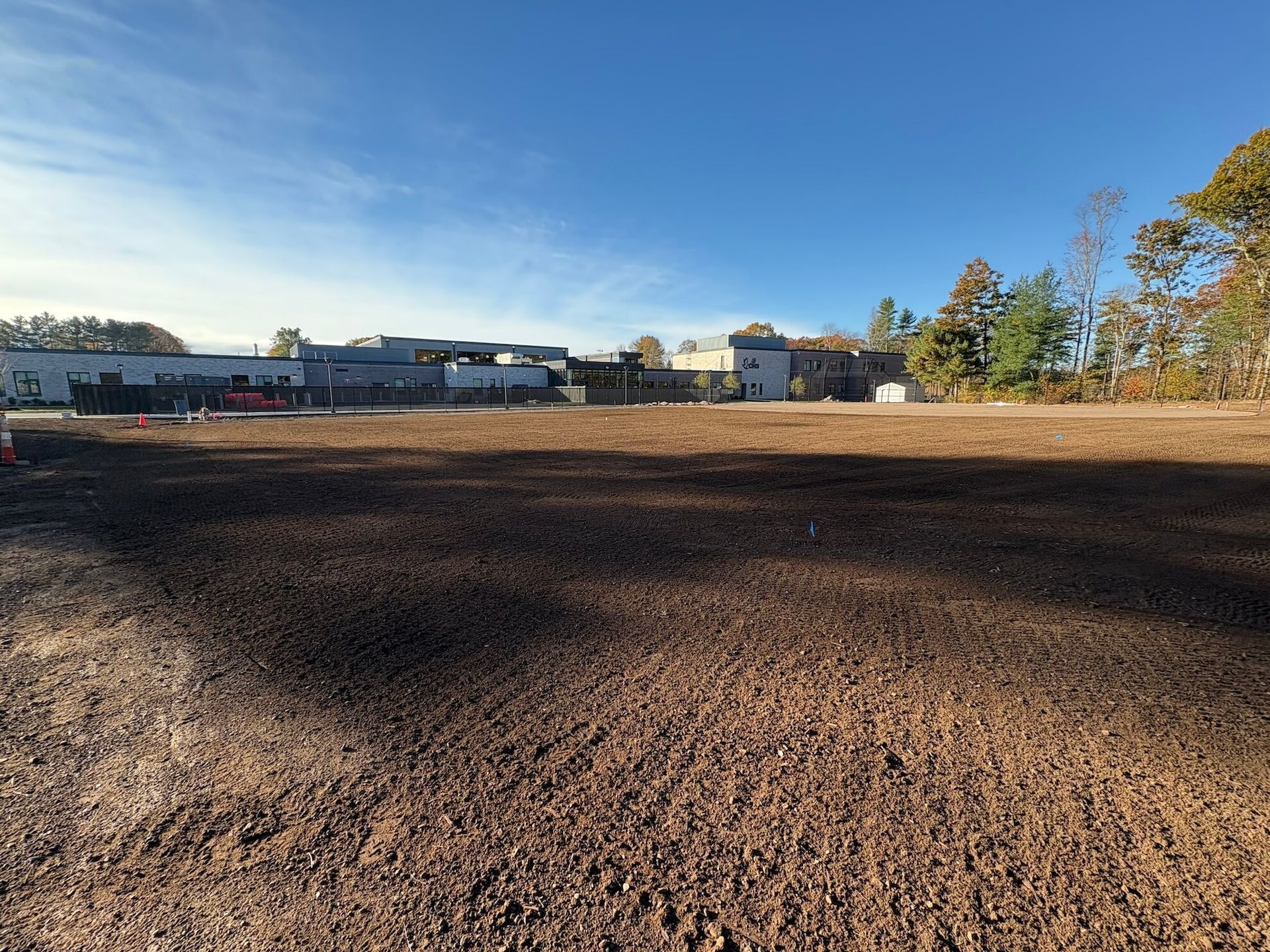 A large dirt lot under a blue sky, with buildings and trees in the distance.