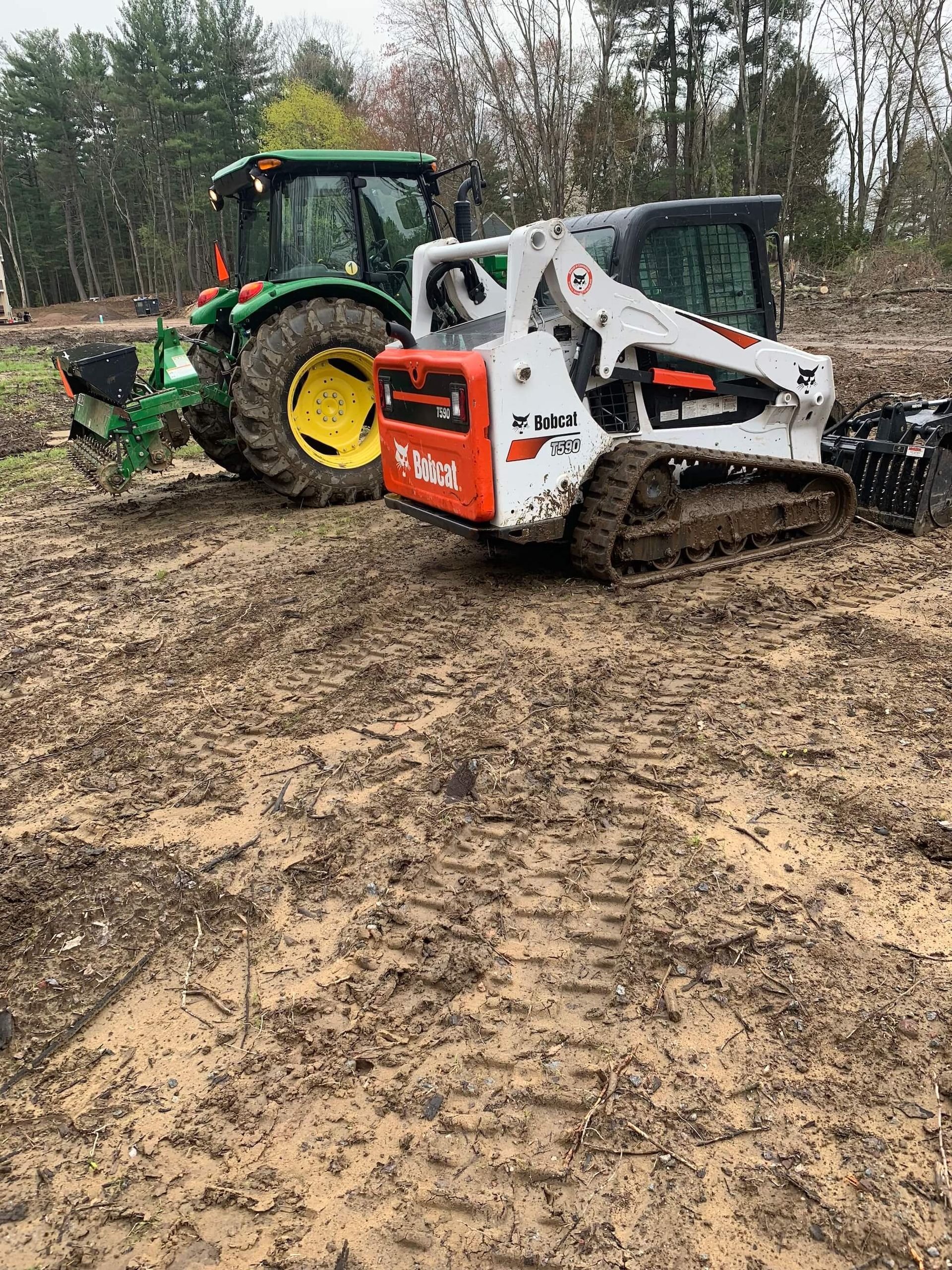 Tractor pulling a white skid steer through a muddy field with trees in the background