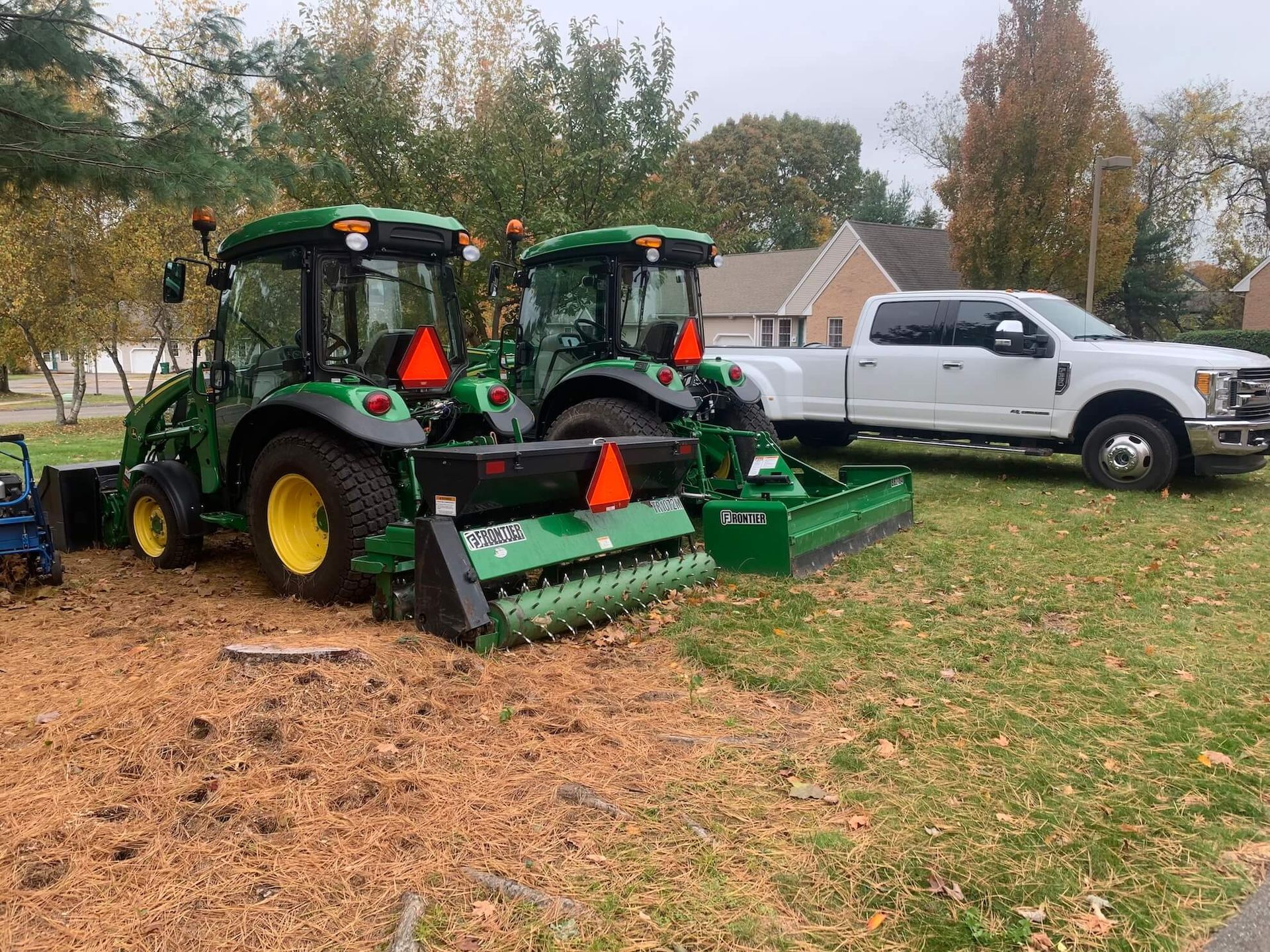 Two green tractors with a mower attachment parked beside a white pickup in a grassy field