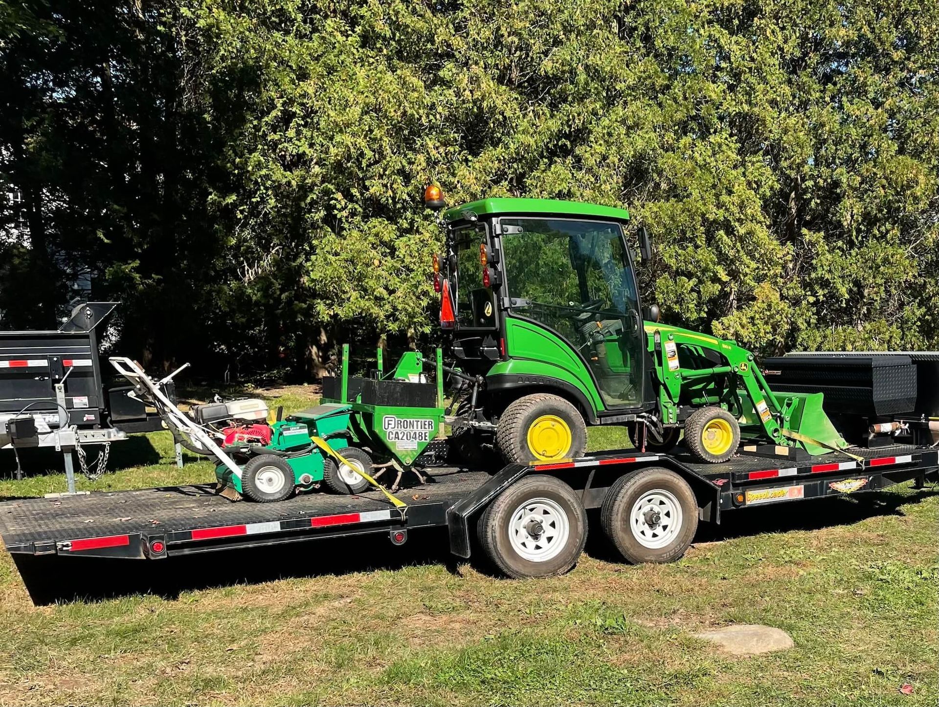 Green tractor on a flatbed trailer in a grassy yard, with trees in the background.