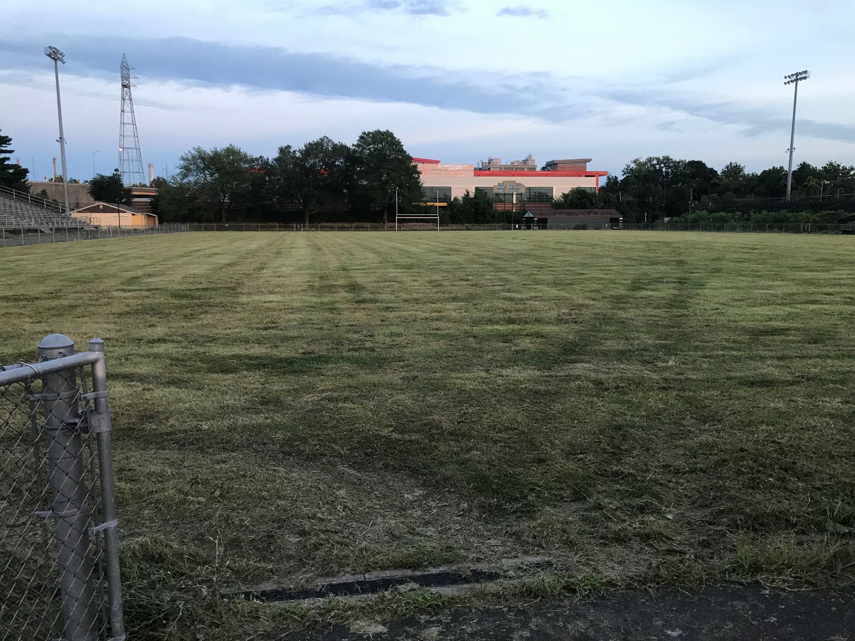 Empty grassy field with trees, a building, and a tall light pole under a cloudy sky
