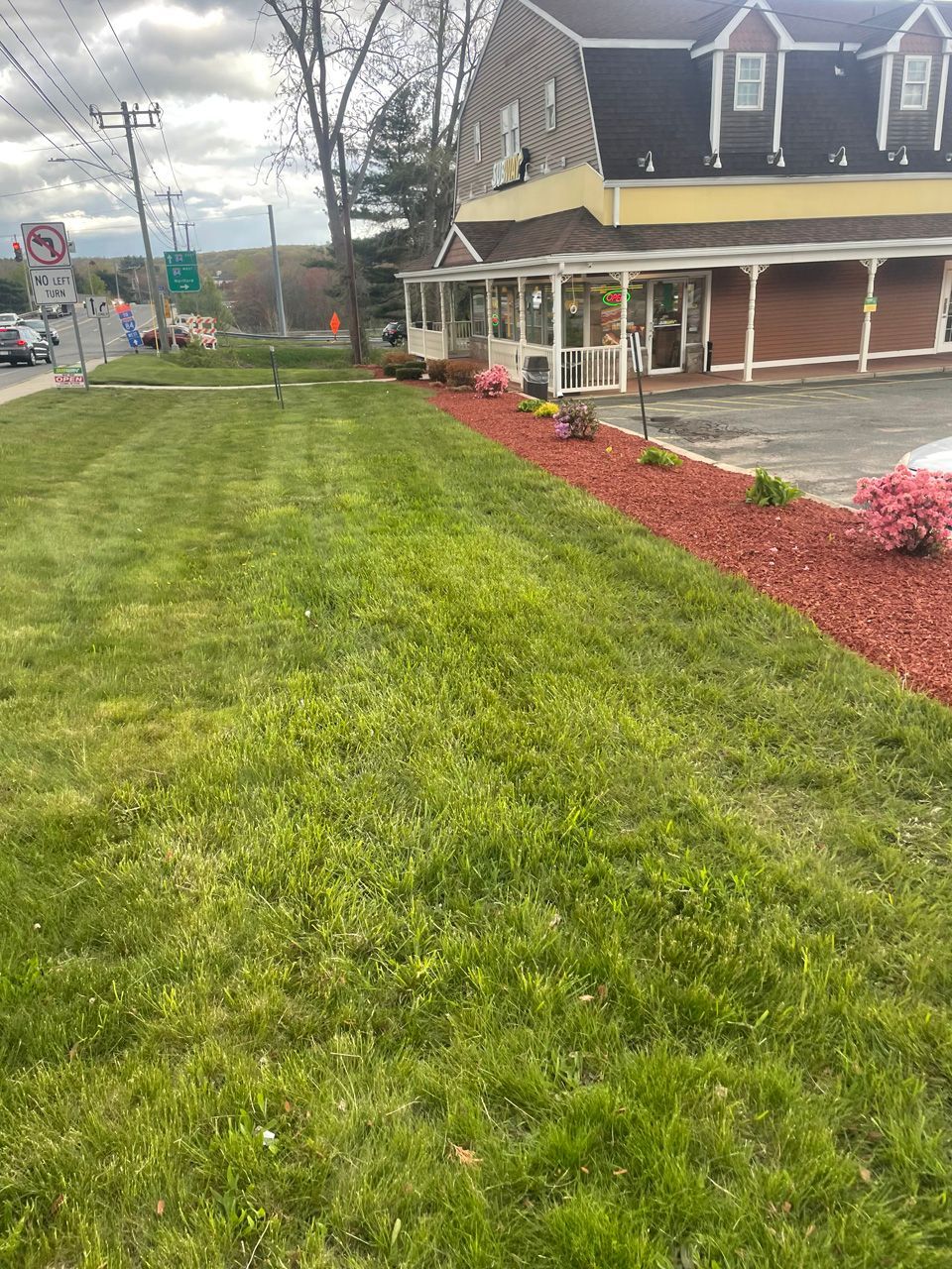 Lawn beside a brick house with porch, red mulch flower beds, and a sidewalk near a street.