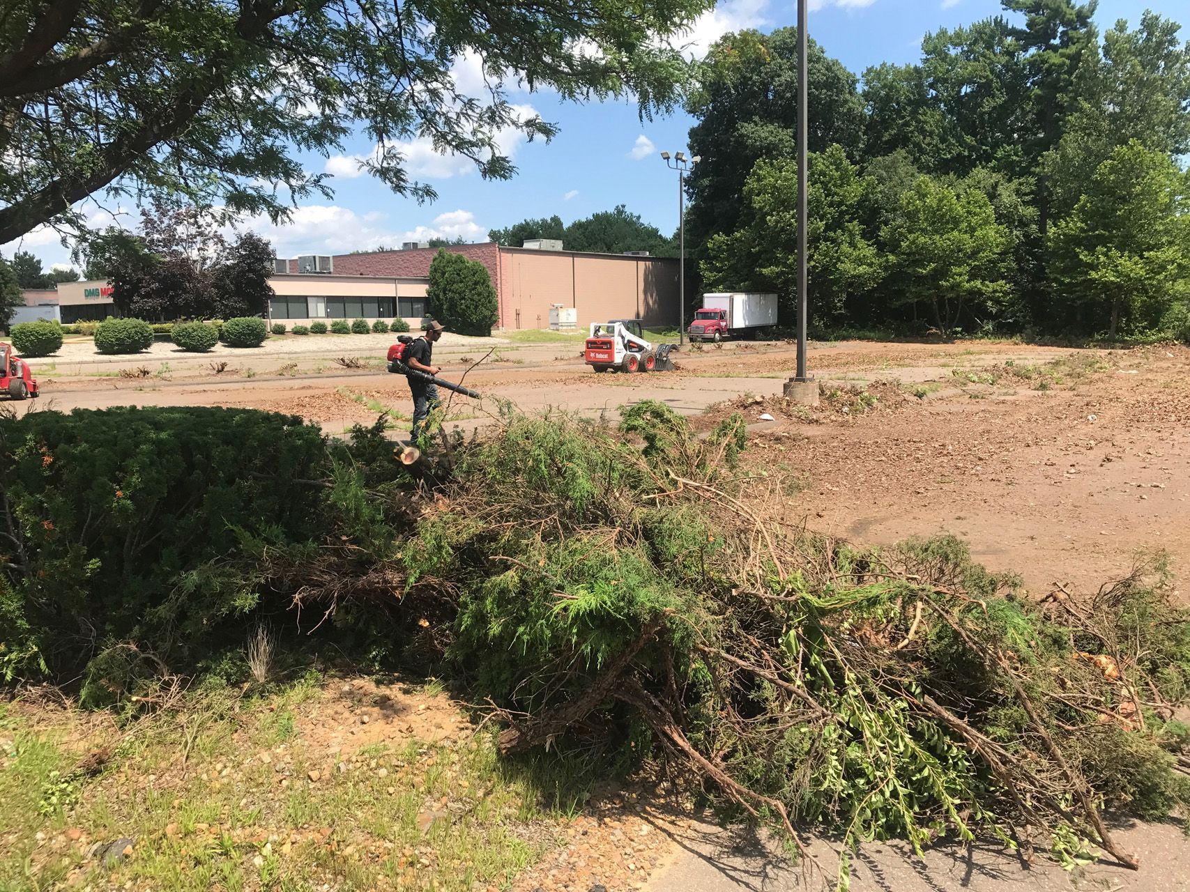 Dirt lot with a person in red near buildings, trees, and a pile of brush under a blue sky