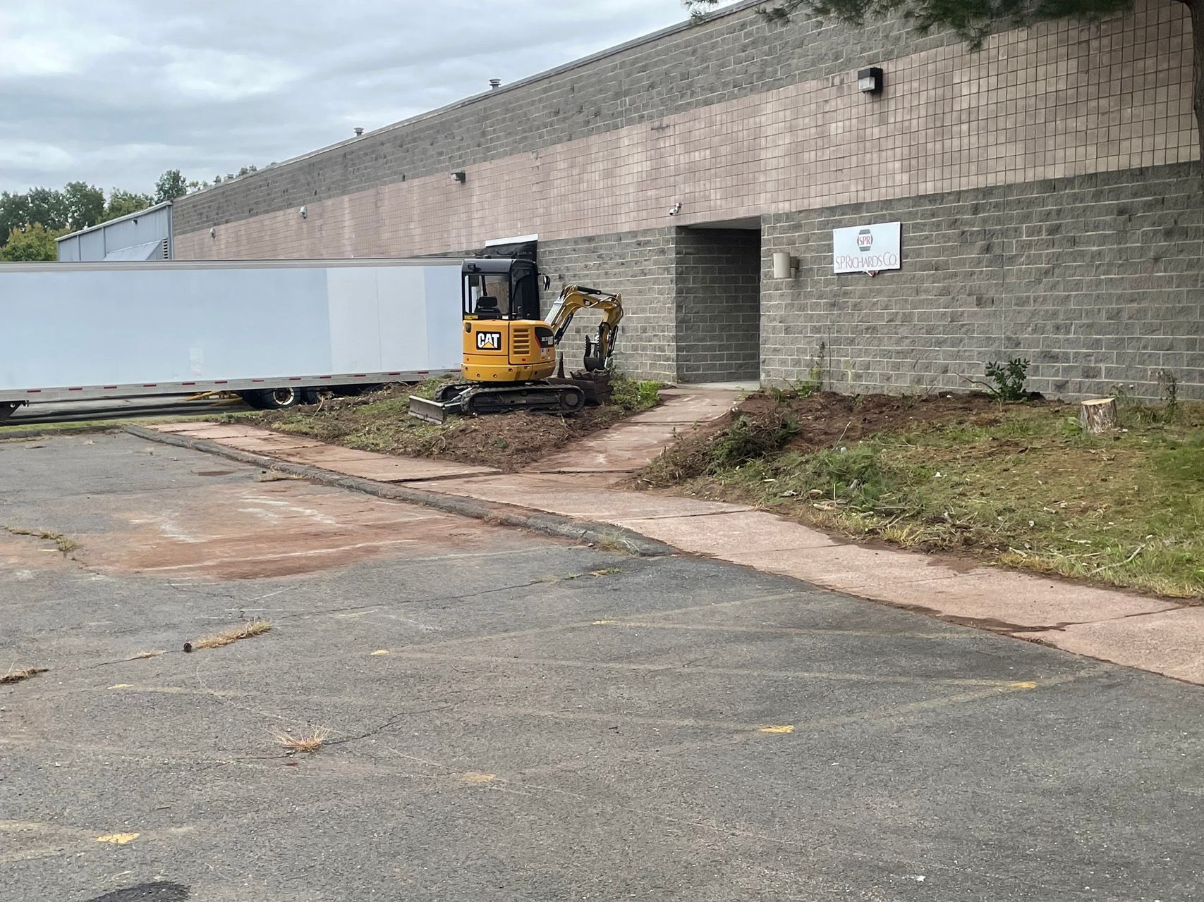 Small yellow excavator beside a brick retaining wall and gate, with a white truck trailer on a lot.