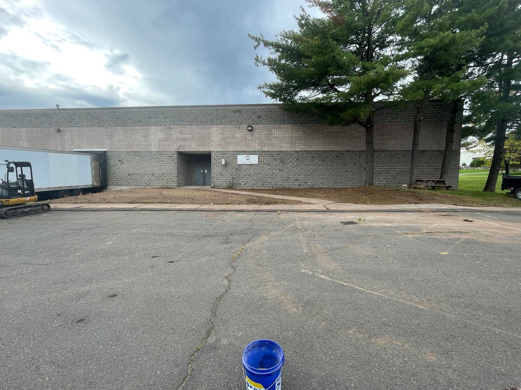 Empty parking lot beside a gray industrial building with a tree and a blue bucket in the foreground