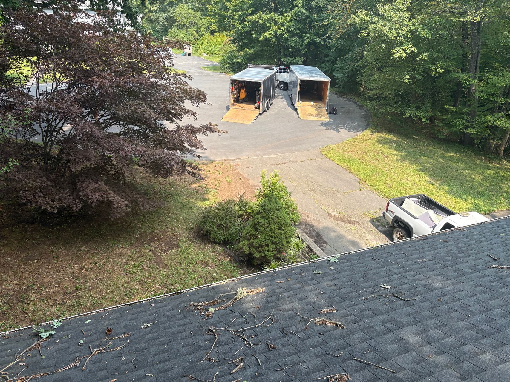 Backyard with a gravel driveway, two trash bins, and a pickup truck near a wooded house.