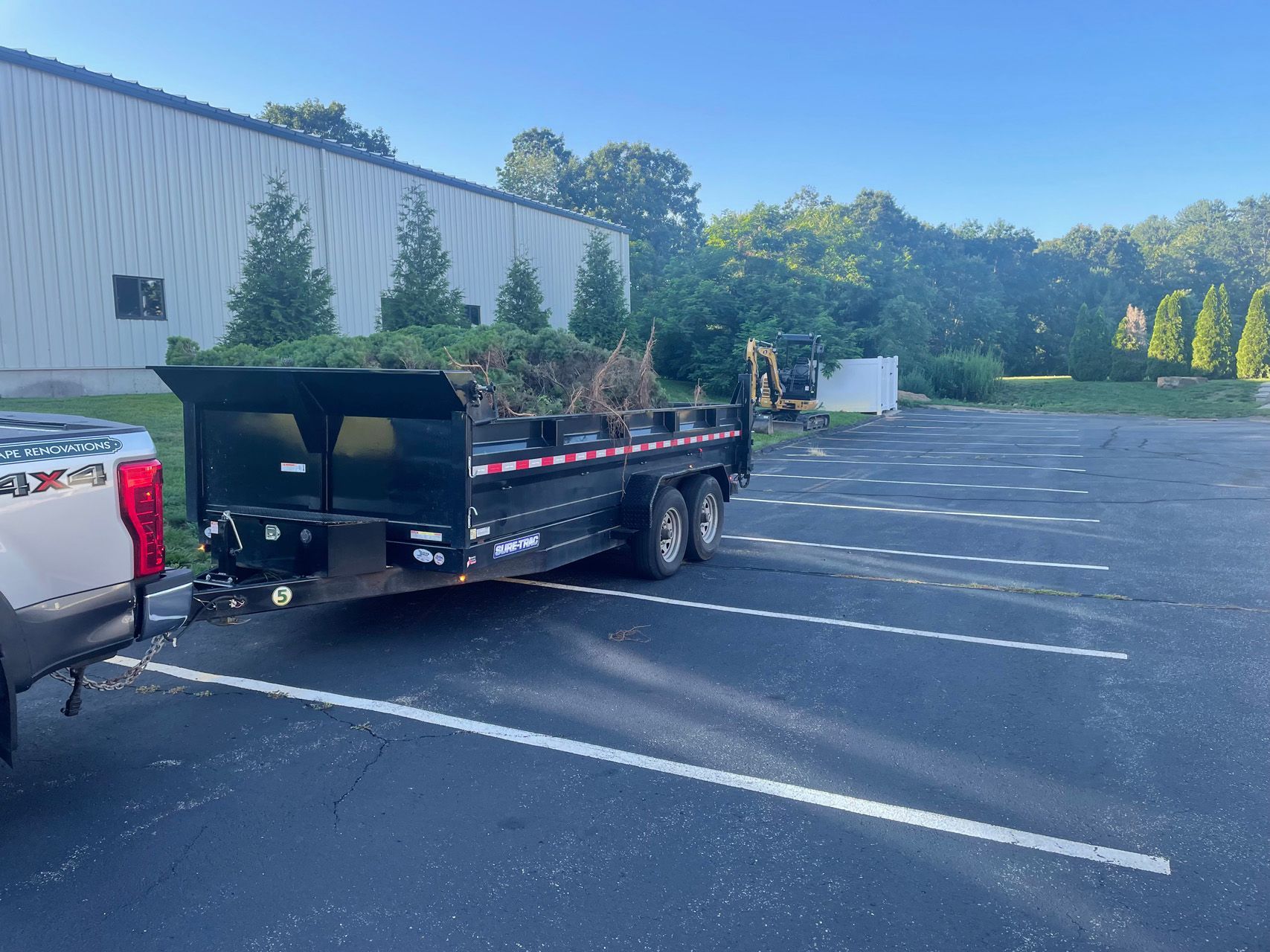 Flatbed trailer hitched to a pickup truck in an empty parking lot beside a white building and trees.