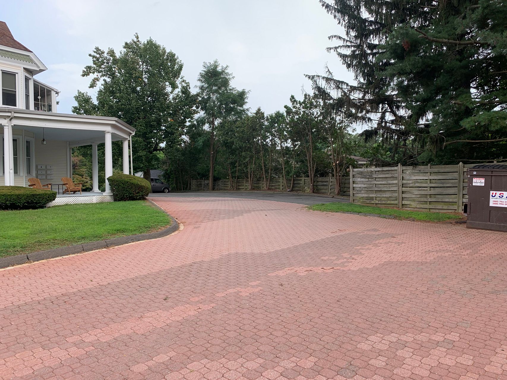 Curved pink brick driveway beside a white house, lawn, trees, and a wooden fence