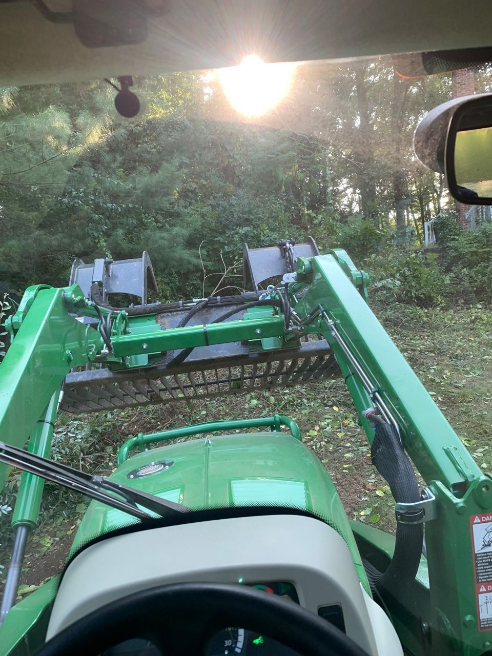 Green tractor front loader in a wooded path, viewed from inside the cab.