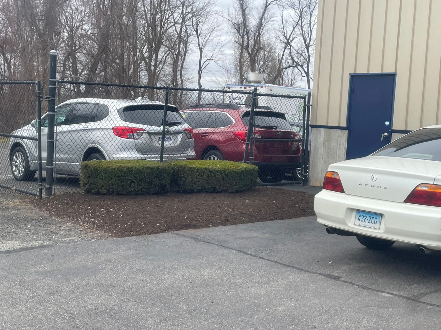 Cars parked beside a building in a snowy lot, with a fence and shrubs in the foreground