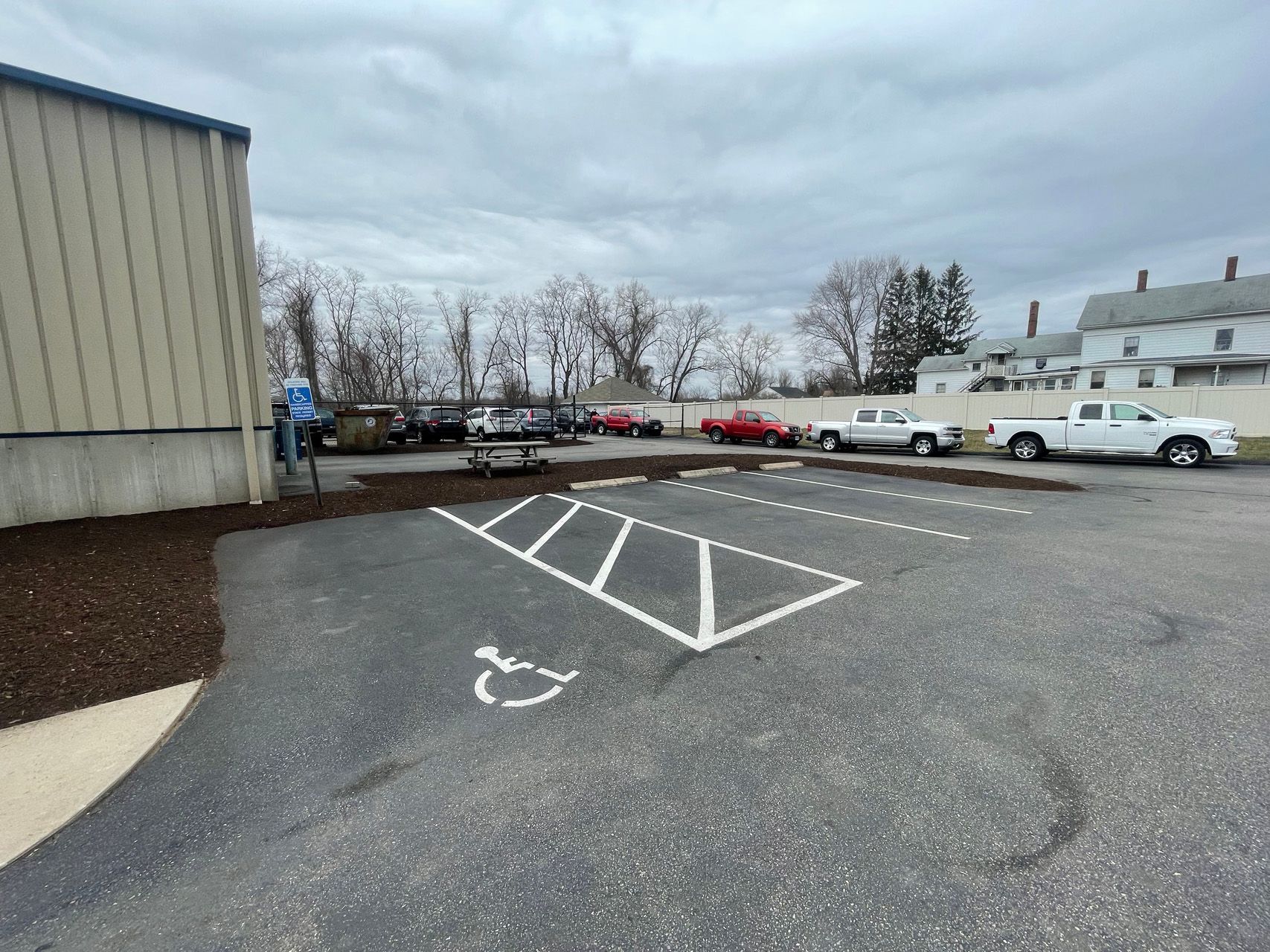 Empty parking lot beside a beige building, with painted handicap lines and several parked vehicles in the distance.