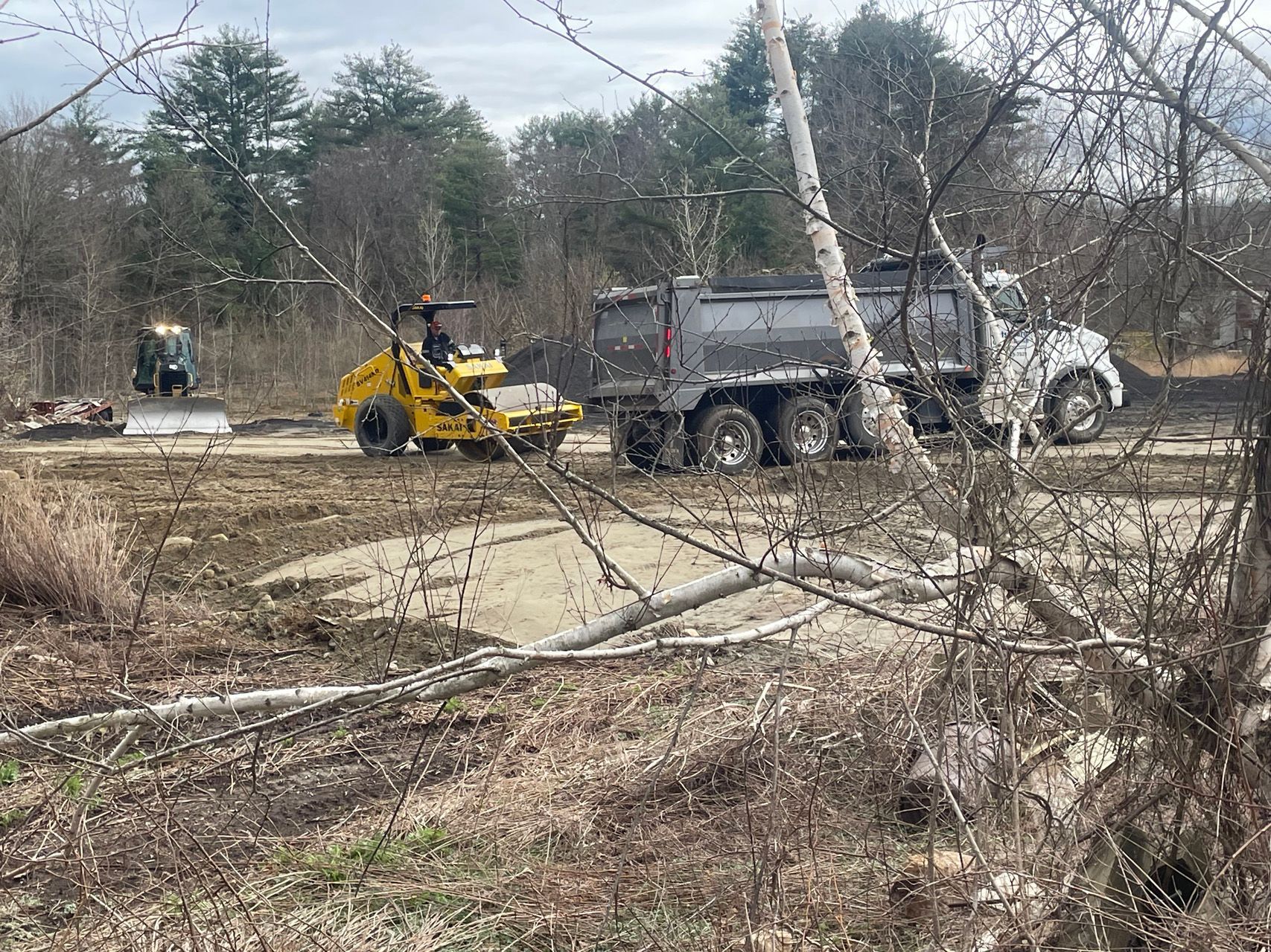 Construction vehicles at a muddy work site, including a yellow loader and gray dump truck, with trees in the background