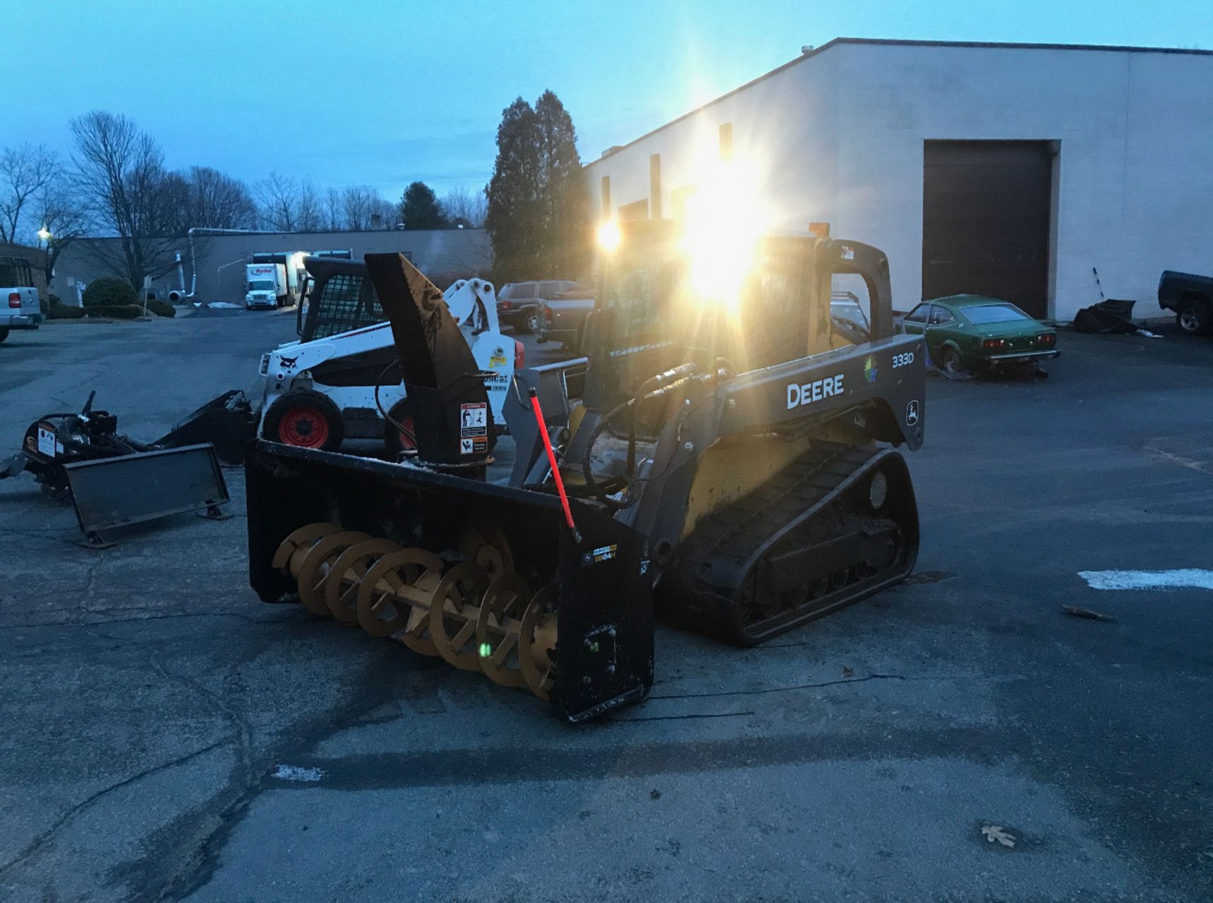 Compact tracked machine with bright headlights parked in a lot at dusk