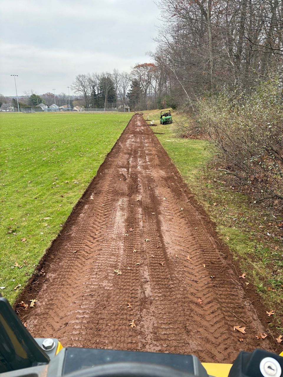 Dirt path through a grassy field toward trees on an overcast day, with part of a vehicle visible in the foreground