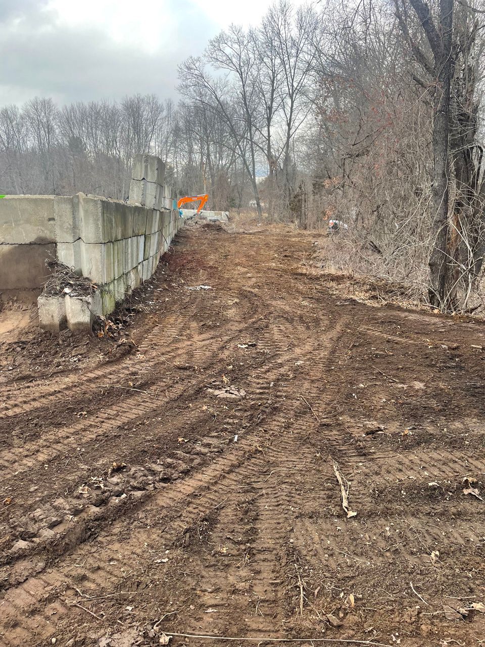 Muddy dirt road under construction beside concrete barriers and leafless trees, with orange caution signs ahead