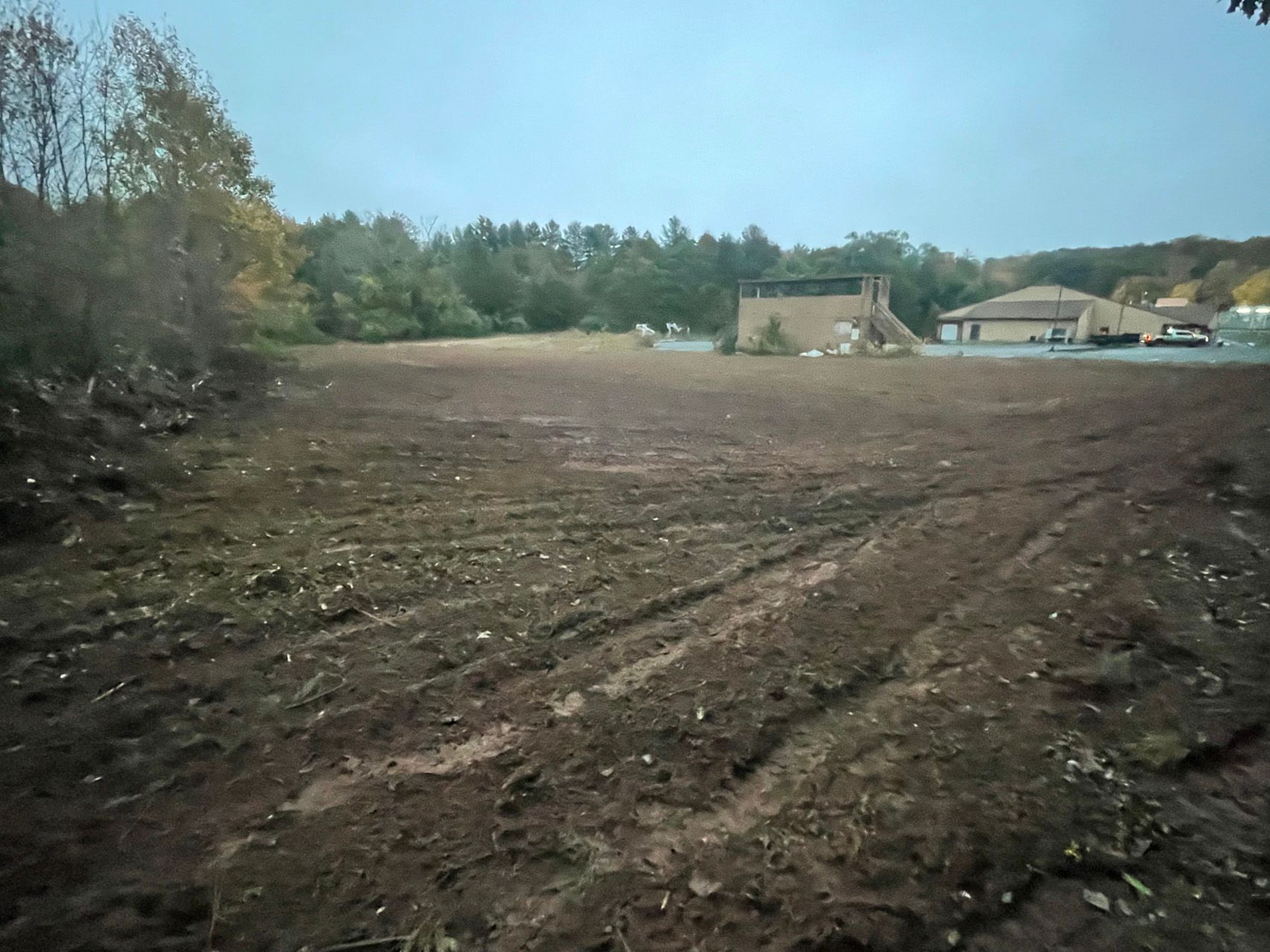 Cleared dirt lot with tire tracks, trees on the left, and houses in the distance under a cloudy sky