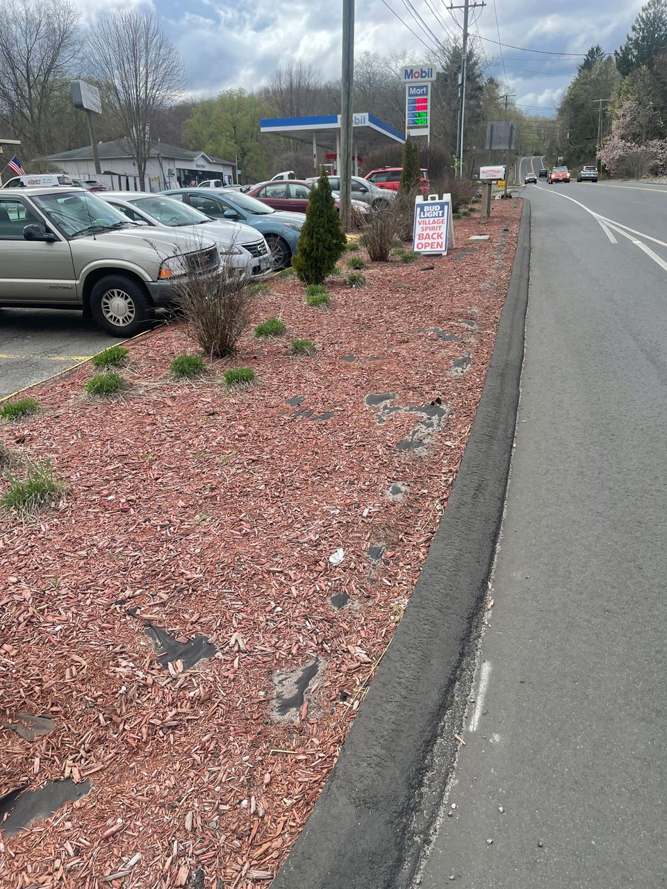 Parking lot with cars beside a road, landscaped with red mulch and small shrubs under cloudy skies.