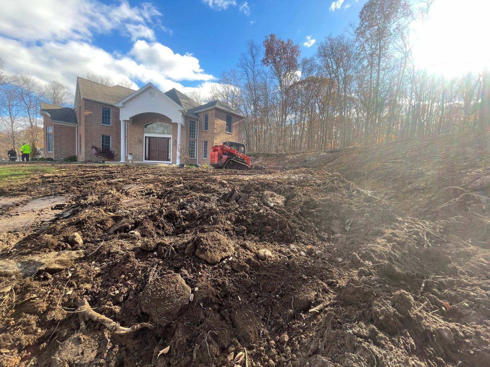 Muddy construction site with a large house, red excavator, and bright sun over leafless trees