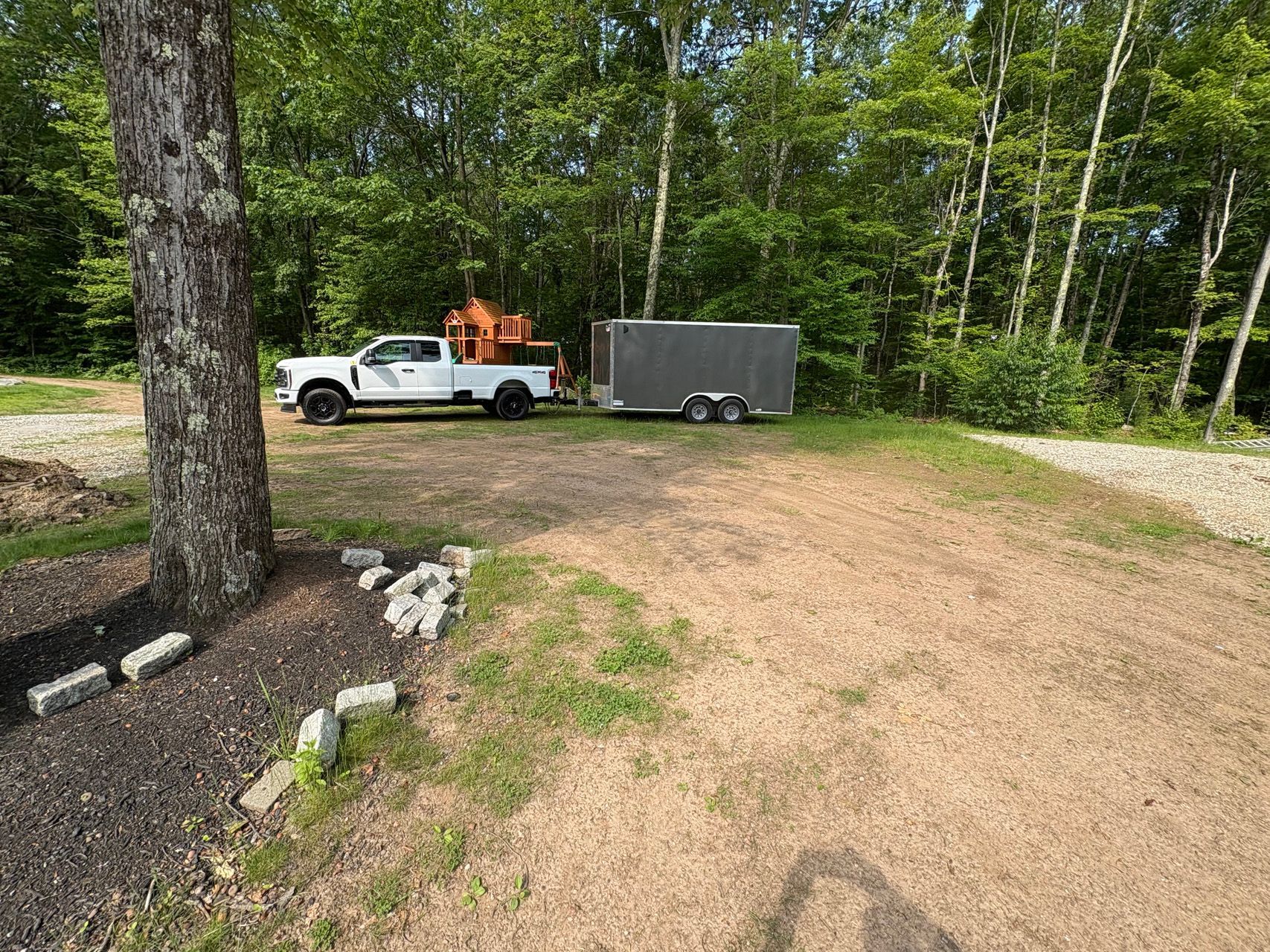 White pickup truck towing a gray trailer on a dirt driveway beside a wooded area