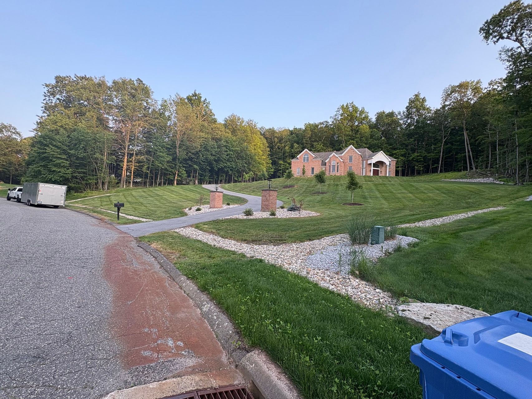 Gravel driveway curving past grassy lots and trees toward a brick house at sunset
