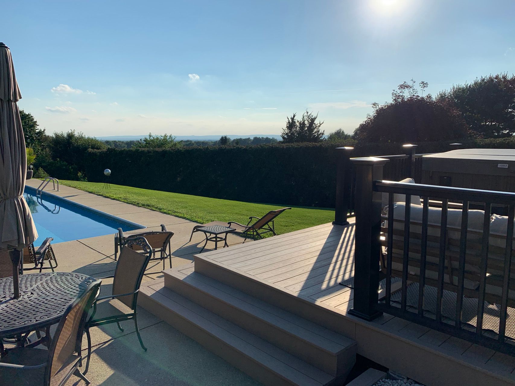 Sunny backyard patio with pool, lounge chairs, stairs, and hedge-lined lawn