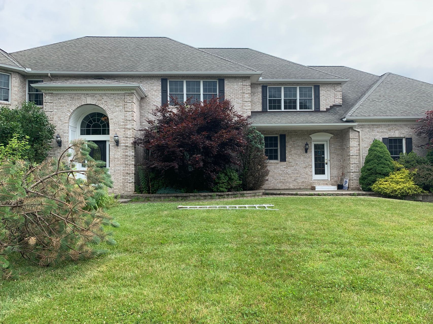 Two-story brick house with a gray roof, arched entry, and a front lawn with shrubs and trees.