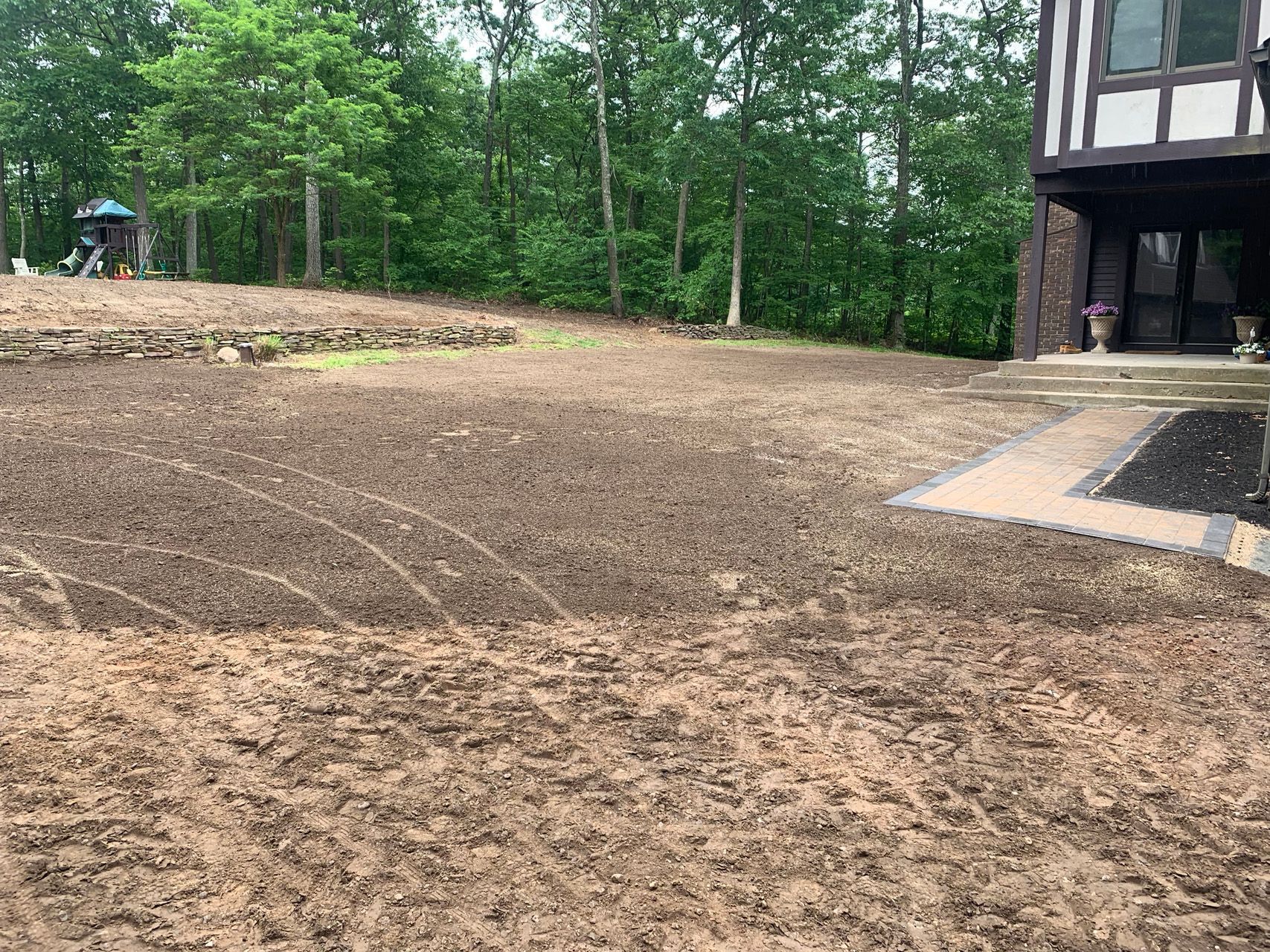 Freshly graded dirt yard beside a raised house, with trees and a small patio area nearby.