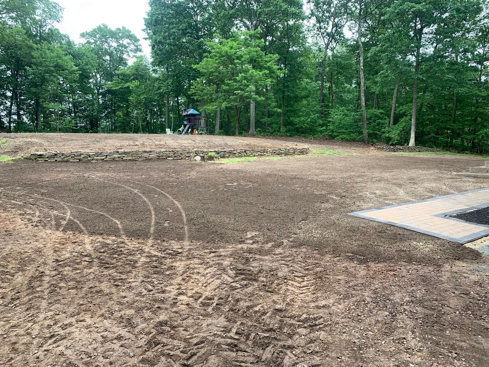 Cleared dirt lot beside a wooded area, with a concrete pad at right and a small person in blue in the distance