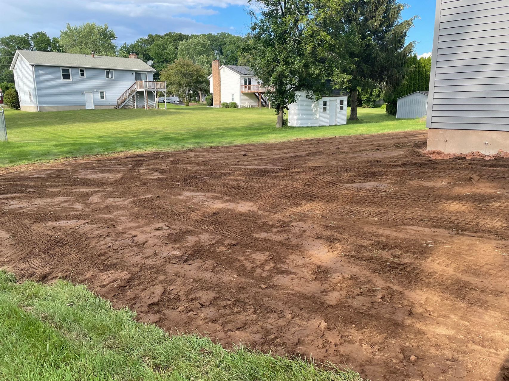 Freshly tilled brown soil beside a gray house in a grassy suburban yard under trees