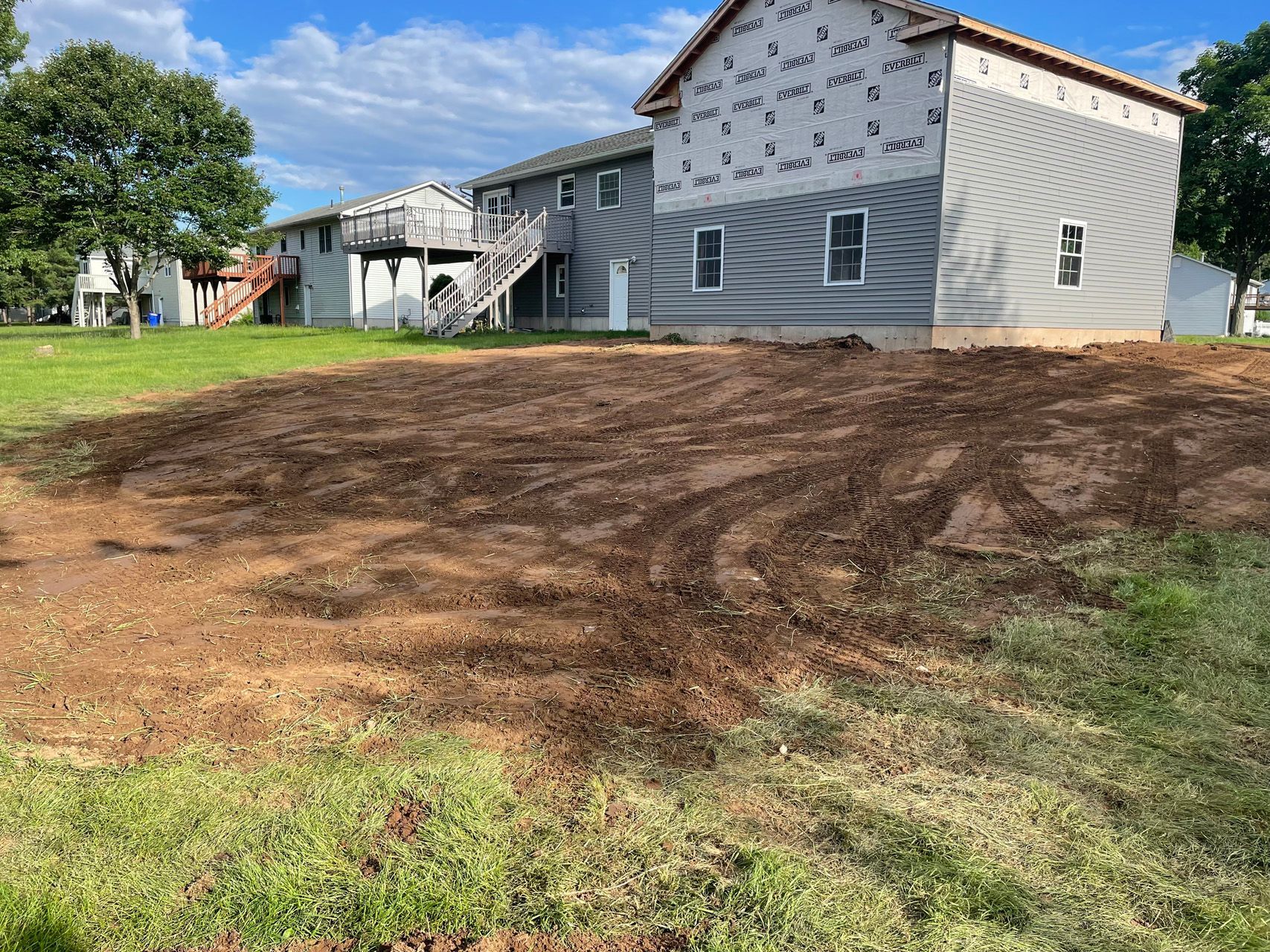 Freshly graded dirt patch beside a white house with gray siding and a grassy yard under a blue sky