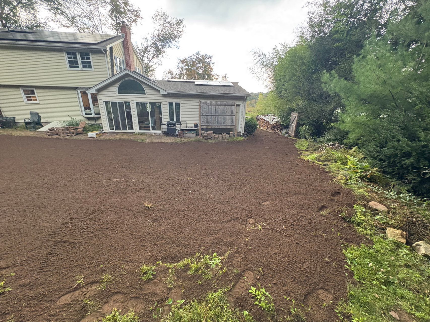 Freshly tilled brown yard beside a house, with trees and a greenhouse-like porch in the background.