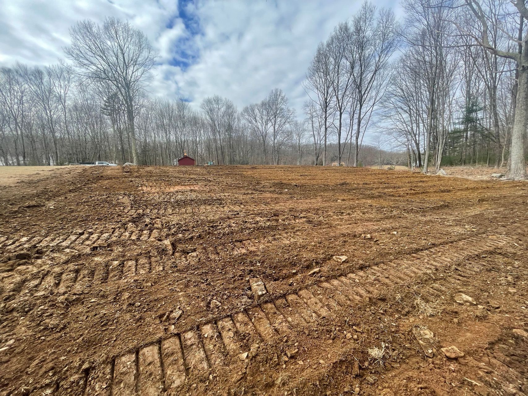 Cleared dirt lot with tire tracks, leafless trees, and a small red building in the distance