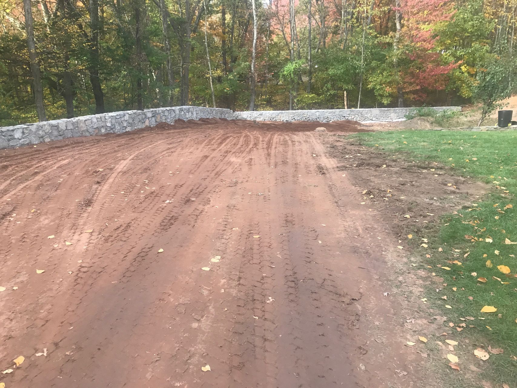 Dirt driveway leading to a stone wall, surrounded by trees with autumn foliage.