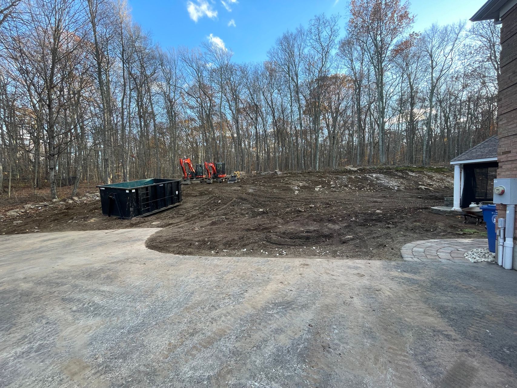 Gravel lot with a dirt mound, red tractor in the background, and leafless trees under a blue sky