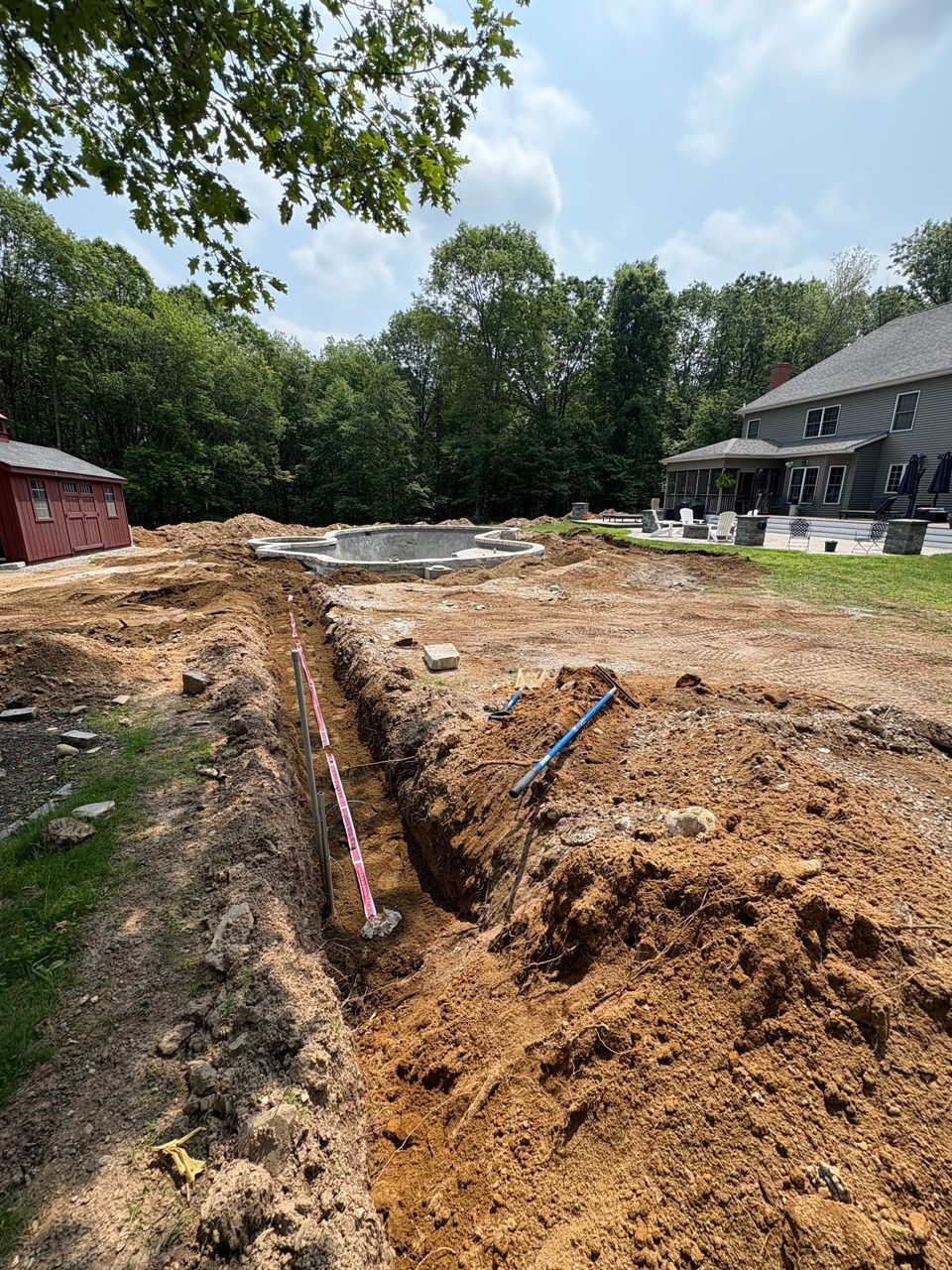 Trenched backyard with exposed pipes, dirt piles, trees, and a house in the background
