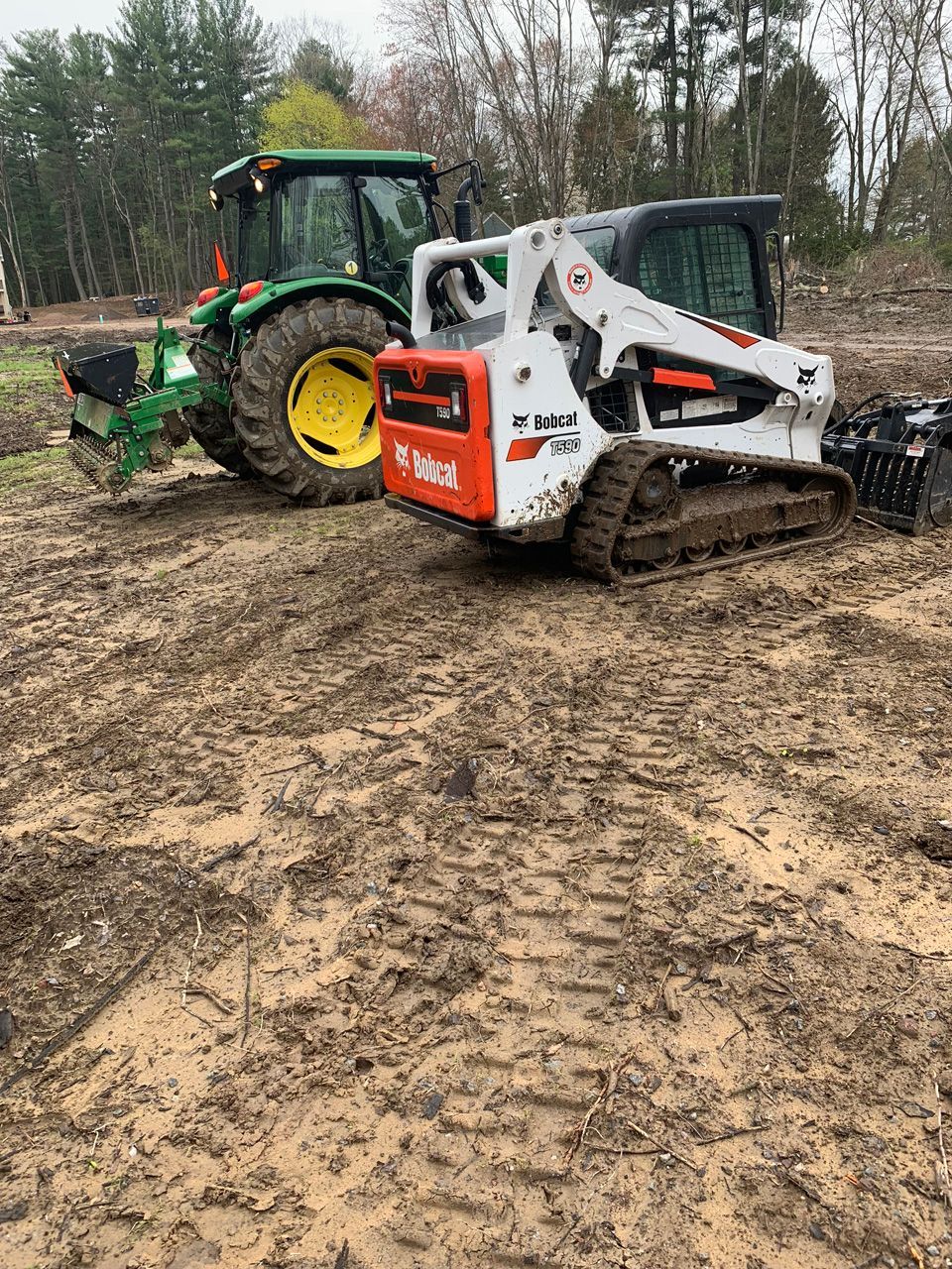 Green tractor and white skid steer working on muddy ground in a wooded area